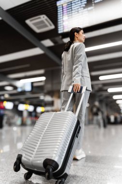 Asyalı kadın kalkış hakkında bilgi almak için havaalanı terminalindeki uçuş çizelgesine bakıyor. Bavullu kız turist uçuş saatine bakıyor. Arkadan görüntüle