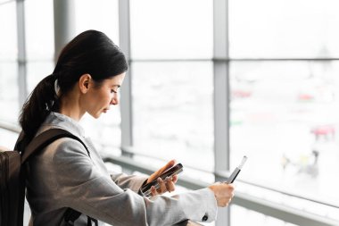 Havaalanındaki turist panoramik pencerenin yanında güvenlik kontrolü için bekliyor. Pasaportu, geçiş kartı ve akıllı telefonu olan bir kadın havaalanı lobisinin penceresinin yanında duruyor ve kalkışı bekliyor.