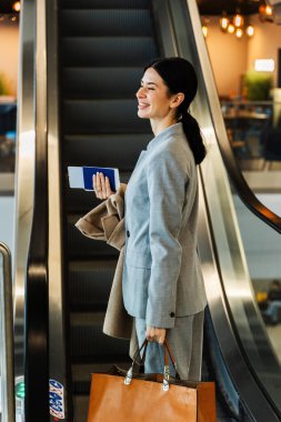 Gri çantalı bir kadın havaalanı terminalinde yürüyen merdivene doğru gidiyor. Havaalanındaki yürüyen merdivene doğru hareket eden turistin arkasından görünüm