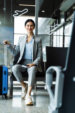 Mavi bavulun sapı kadının elinde ve havaalanı terminaline bakıyor. İş kadını havaalanında oturuyor ve seyahatine başlamayı bekliyor.