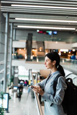 Havaalanı lobisinde akıllı telefonu ve pasaportu olan çekici bir kadın. Havaalanı terminalinde yürüyen merdivenin yanında belgelerle bekleyen gezgin 2024 yaz tatilinin başlamasını bekliyor.