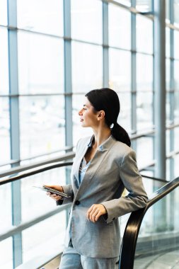 Havaalanı terminalinde yürüyen merdivenin yanında belgelerle bekleyen gezgin 2024 yaz tatilinin başlamasını bekliyor. Havaalanı lobisindeki akıllı telefondan bakan kadın..