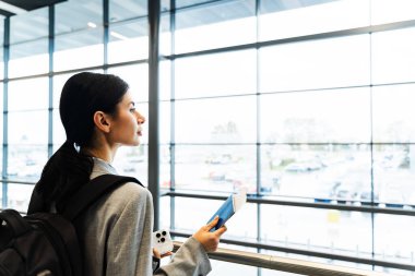 Havaalanı salonunda akıllı telefon, belge ve sırt çantalı bir turist panoramik pencereden bakıyor. Uçuşun 2024 'te başlamasını bekleyen gülümseyen beyaz kadın.
