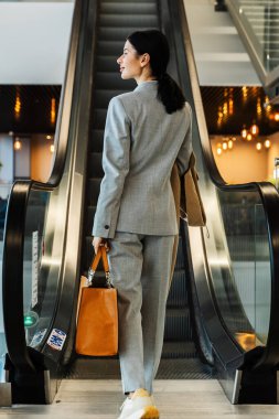 Gri çantalı bir kadın havaalanı terminalinde yürüyen merdivene doğru gidiyor. Havaalanındaki yürüyen merdivene doğru hareket eden turistin arkasından görünüm