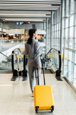 Seyahat ve tatil konsepti. Gezgin kız 2024 yazında yeni bir gezi bekliyor. Sarı valizli Asyalı kadın havaalanı terminalindeki yürüyen merdivene yürüyor.