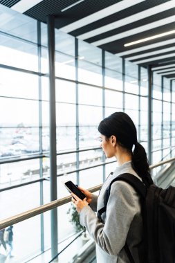 Havaalanı terminalinde akıllı telefonunu kullanarak uçuş numarasını kontrol eden bir kız. Seyahat ve tatil kavramı