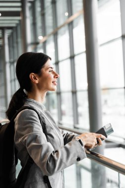 Havaalanındaki turist panoramik pencerenin yanında güvenlik kontrolü için bekliyor. Pasaportu, geçiş kartı ve akıllı telefonu olan bir kadın havaalanı lobisinin penceresinin yanında duruyor ve kalkışı bekliyor.
