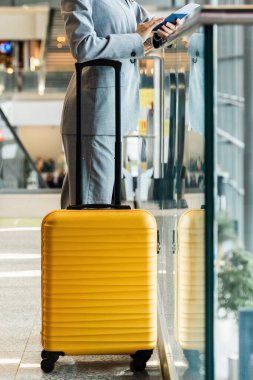Panorama penceresinden bakan valizli bir iş kadını tatil için uçağa binmeyi bekliyor. Havaalanı terminalinde iş elbisesi giymiş, sarı bavullu, camdan bakan Asyalı bir kız.
