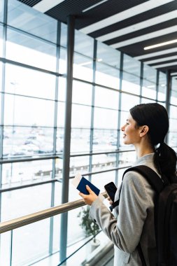 Havaalanı salonunda akıllı telefon, belge ve sırt çantalı bir turist panoramik pencereden bakıyor. Uçuşun 2024 'te başlamasını bekleyen gülümseyen beyaz kadın.