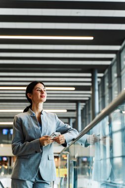 Havaalanı salonunda akıllı telefon, belge ve sırt çantalı bir turist panoramik pencereden bakıyor. Uçuşun 2024 'te başlamasını bekleyen gülümseyen beyaz kadın.
