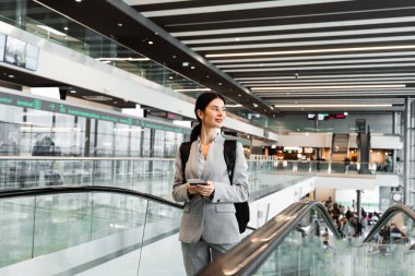 Siyah sırt çantalı Asyalı kız havaalanı terminalinde yürüyen merdivende yürüyor. Seyahat ve tatil konsepti. İş kadını 2024 yazında yeni bir gezi bekliyor.