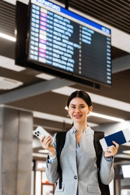 Pasaportlu ve akıllı telefonlu heyecanlı öğrenci kalkış kurulu arka planında. Genç Asyalı kız havaalanı lobisinde kalkış için bekliyor.