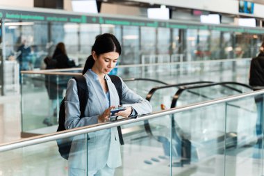 Havaalanı terminalinde yürüyen merdivenin yanında belgelerle bekleyen gezgin 2024 yaz tatilinin başlamasını bekliyor. Havaalanı lobisindeki akıllı telefondan bakan kadın.
