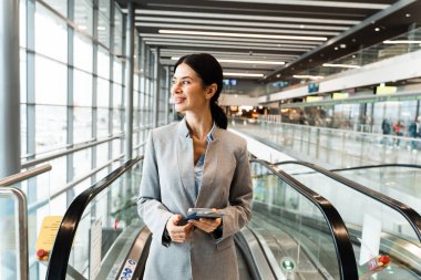 Havaalanı terminalinde yürüyen merdivenin yanında belgelerle bekleyen gezgin 2024 yaz tatilinin başlamasını bekliyor. Havaalanı lobisindeki akıllı telefondan bakan kadın..
