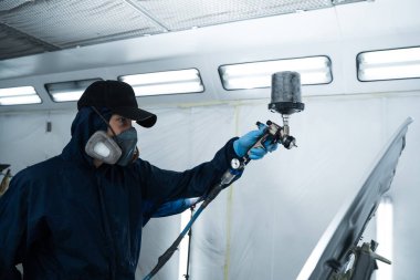 Araç bakım servisindeki tamirci koyu renk sprey ve boya tabancası kullanarak arabaya boya sürüyor. Koruyucu eldiven giyen profesyonel işçi otomobili boyuyor