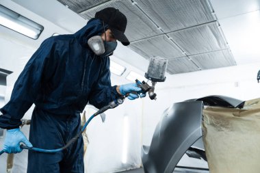 Araç bakım servisindeki tamirci koyu renk sprey ve boya tabancası kullanarak arabaya boya sürüyor. Koruyucu eldiven giyen profesyonel işçi otomobili boyuyor