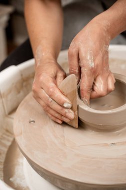 Kız, ahşap bir kazıyıcı ve çömlek tekeriyle çanak çömleğin pürüzsüz kenarlarını yapıyor. Kil bir ürünün kenarlarını tahta bir kazıyıcıyla kesiyor.