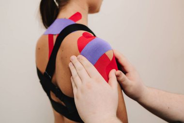 Atlet performansını arttırmak için boynunda ve omuzlarında ince, esnek bantlar olan genç bir kadın. Doktor kasları çalıştırmak için hastanın omzuna kineziyoloji bantları takıyor.