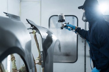 Araba boyacısı otomobil boyuyor. Araç bakım servisindeki tamirci koyu renk sprey ve boya tabancası kullanarak arabanın üzerine boya saçıyor.