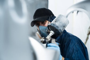 Araç bakım servisindeki tamirci koyu renk sprey ve boya tabancası kullanarak arabaya boya sürüyor. Koruyucu eldiven giyen profesyonel işçi otomobili boyuyor