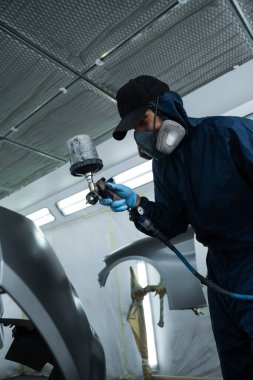 Araç bakım servisindeki tamirci koyu renk sprey ve boya tabancası kullanarak arabaya boya sürüyor. Koruyucu eldiven giyen profesyonel işçi otomobili boyuyor