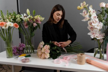 Teslimat için kurdeleyle pembe güllerin çiçek bileşimini bağlar. Taze çiçekler. Kadın çiçekçi çiçek atölyesindeki masada bir buket gül yapıyor.
