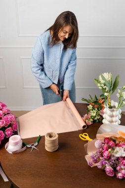 Çiçekçi çiçek kompozisyonunu el işi kağıdına sarar. Çiçekçi çiçekçi dükkanındaki ahşap masada çiçek buketi için ambalaj kağıdı hazırlıyor.