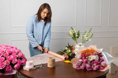 Çiçekçi çiçekçide ahşap masanın üzerinde çiçek buketi için ambalaj kağıdı hazırlıyor. Kadın işçi çiçeklerin bileşimini el sanatları kağıdına sarar