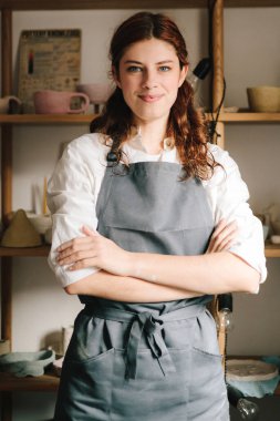 Küçük işletmeleri başarılı bir zanaatkar kadın yönetiyor. Onun çömlekçi stüdyosunun arka planında toprak malzemeleriyle dolu raflarla mutlu genç çömlekçi duruyor ve gülümsüyor.