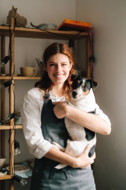 Çömlek atölyesinin arka planında saklama eşyalarıyla dolu raflarla gülümseyen genç ve mutlu bir çömlekçi. Küçük işletmeleri başarılı bir zanaatkar kadın yönetiyor.