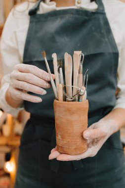 Siyah önlüklü Potter seramik kupada çömlek yontmak için bir sürü alet tutuyor. Seramikçiler için kil işçiliği araçları