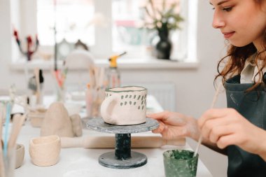 Sanatçı yaratıcı atölyede el yapımı seramik kil bardak süsleme ve boyama yapıyor. Ressam, toprak eşyasına eşsiz tasarımlar çiziyor ve ekliyor