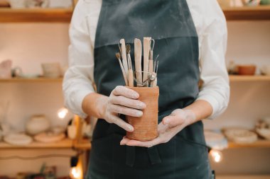 Seramikçiler için kil işçiliği aletleri. Siyah önlüklü Potter seramik bardakta çömlek yontmak için bir sürü enstrüman tutuyor.