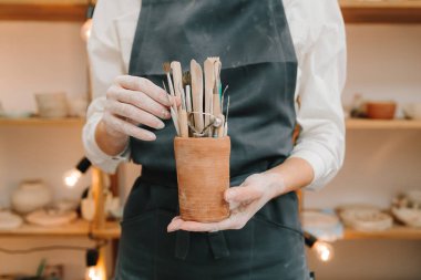 Seramikçiler için kil işçiliği aletleri. Siyah önlüklü Potter seramik bardakta çömlek yontmak için bir sürü enstrüman tutuyor.