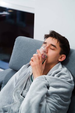 Evde öksürük tedavisi için nebulizör maskesiyle hava solumak. Hasta adam nebulizatörle solunum tedavisi görüyor.