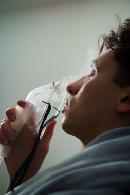 Battaniyeye sarılı hasta adam öksürük tedavisi için nebulizatörle solunum yapıyor. Hasta evde tedavi için astım spreyi kullanıyor.