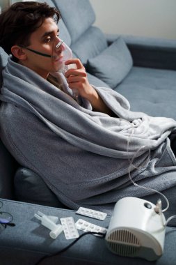 Battaniyeye sarılı hasta adam öksürük tedavisi için nebulizatörle solunum yapıyor. Hasta evde tedavi için astım spreyi kullanıyor.