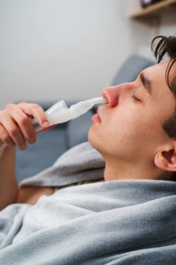 Evde nebulizörle burun solumuş. Astım krizi geçirmiş genç bir adam solunum cihazıyla nefes alıyor.