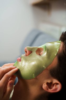 Kırışıklık önleme prosedürü ve evde yeşil maske. Adam yaşlanmayı önleyen yüz maskesi takıyor.