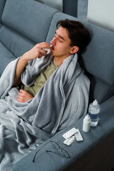 Adam burun spreyi kullanıyor ve hastalık tedavisi için ilaç ve ilaç alıyor. Adam üşüyor ve yatakta uzanıyor ve burnunu burun spreyiyle siliyor.