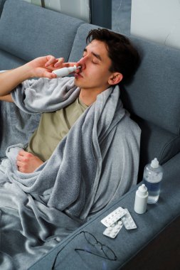 Adam burun spreyi kullanıyor ve hastalık tedavisi için ilaç ve ilaç alıyor. Adam üşüyor ve yatakta uzanıyor ve burnunu burun spreyiyle siliyor.