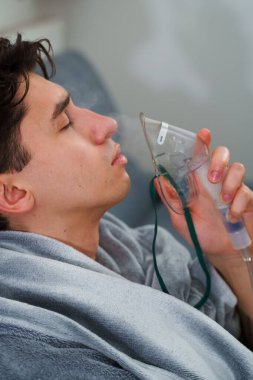 Evde öksürük tedavisi için nebulizör maskesiyle hava solumak. Hasta adam nebulizatörle solunum tedavisi görüyor.