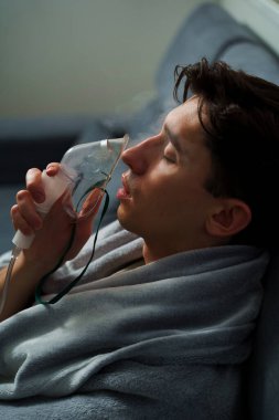 Battaniyeye sarılı hasta adam öksürük tedavisi için nebulizatörle solunum yapıyor. Hasta evde tedavi için astım spreyi kullanıyor.