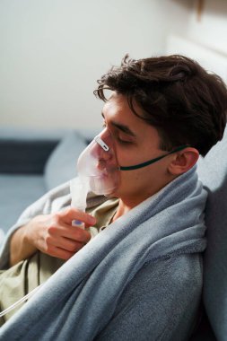 Battaniyeye sarılı hasta adam öksürük tedavisi için nebulizatörle solunum yapıyor. Hasta evde tedavi için astım spreyi kullanıyor.