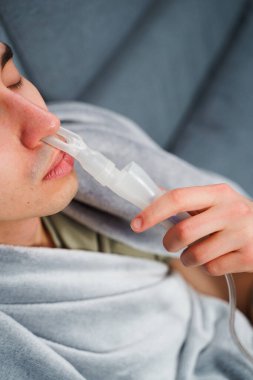 Evde nebulizörle burun solumuş. Astım krizi geçirmiş genç bir adam solunum cihazıyla nefes alıyor.