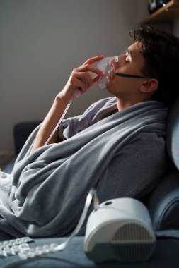 Öksürük tedavisi için oksijen maskesi yutan bir adam. Evde hastalık tedavisi için kompresör nebulizörü kullanılıyor.