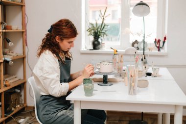 Sanatçı yaratıcı atölyede el yapımı seramik kil bardak süsleme ve boyama yapıyor. Ressam, toprak eşyasına eşsiz tasarımlar çiziyor ve ekliyor