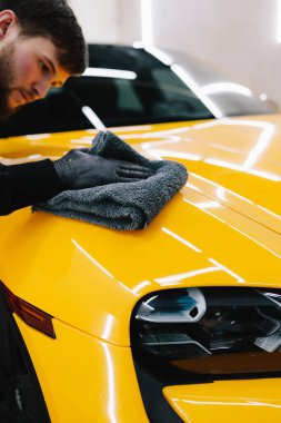 Temizlikçi, sarı bir arabanın vücudunu mikrofiber bezle siler. Tamirci arabayı temizlemek için gri bir mikrofiber bez kullanıyor.