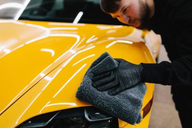 Tamirci, arabayı silmek için gri bir mikrofiber bez kullanıyor. Temizlikçi, sarı bir arabanın vücudunu mikrofiber bezle siler.