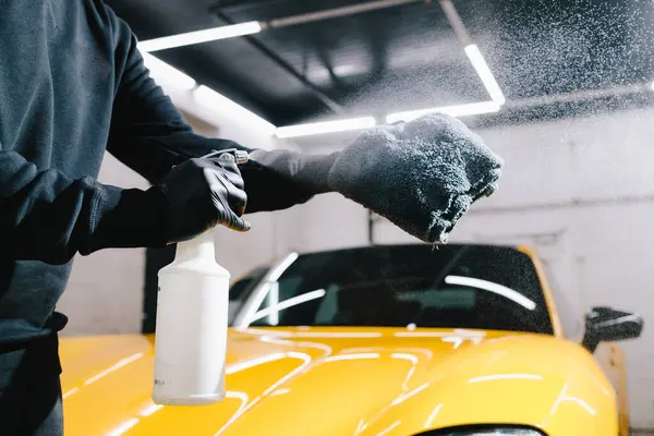 Otomobil servisinde mikrofiberin üzerine mum spreyli araba temizliği. Adam detaylı işlem sırasında araba seramik cilalama için mikrofiber kumaşa balmumu ürünü püskürtür.
